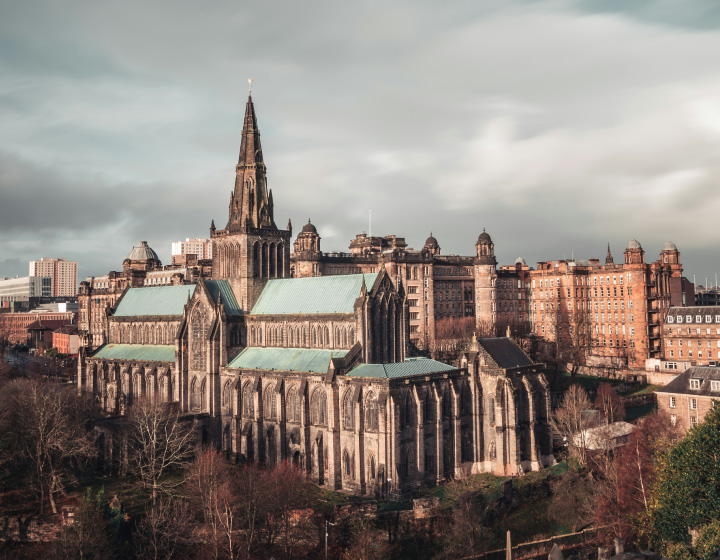 Aerial view of a historic cathedral with a tall central spire and green roof, surrounded by city buildings under a cloudy sky.