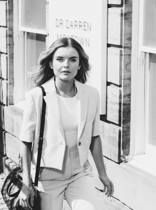 Black and white image of a woman walking outside a clinic building, carrying a bag and dressed in smart, professional attire