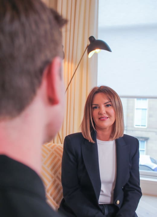 Woman in a blazer speaking during a consultation while a male practitioner is seen in the foreground in a modern clinic setting.