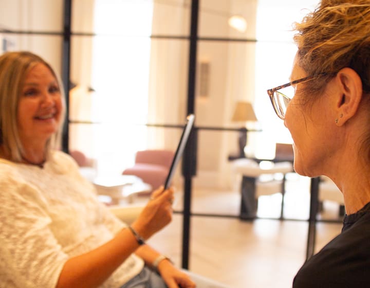Dr. Sharon Ritchie smiling during a patient consultation as the patient looks into a handheld mirror in a bright, modern clinic setting.
