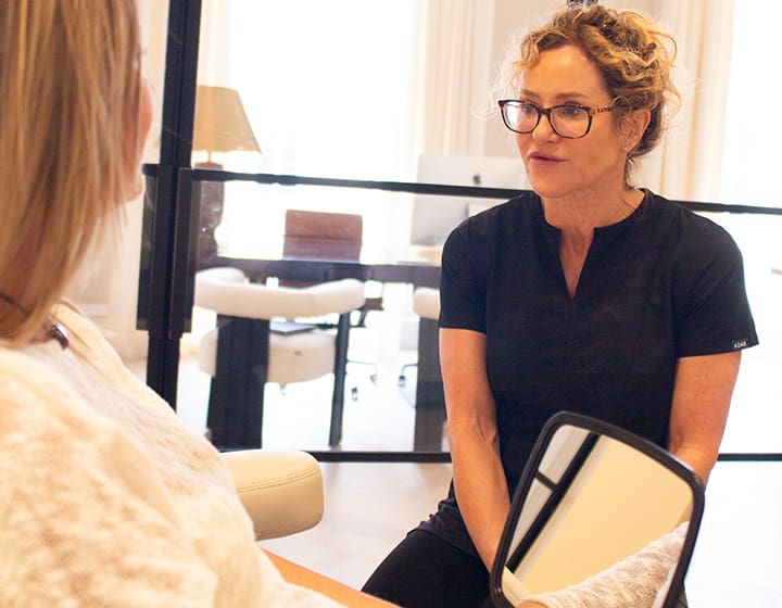 Dr. Sharon Ritchie consulting with a patient in a modern clinic, holding a mirror while discussing facial treatment results.